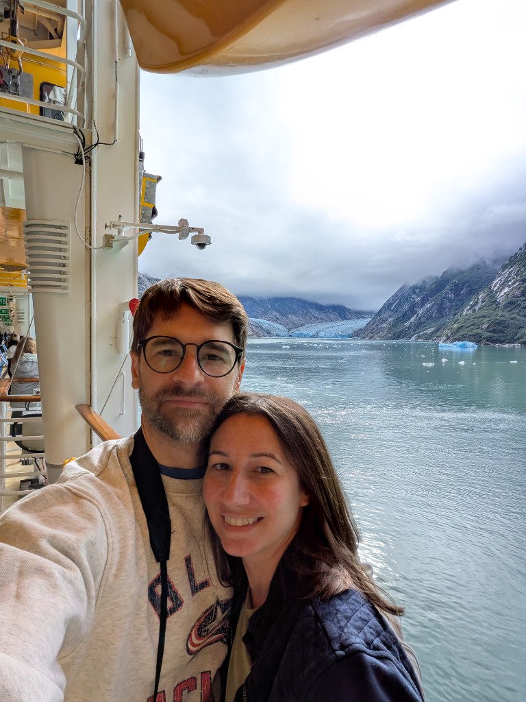 John and Alana Van Fossen smiling together on a ship deck, with icebergs and a glacier in the distance.